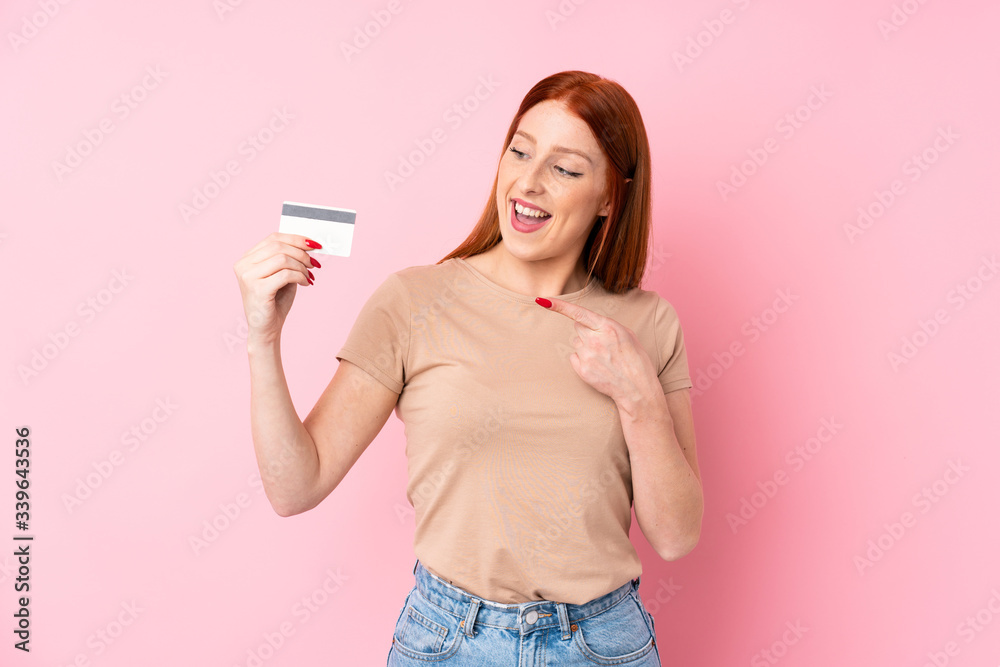 Young redhead woman over isolated pink background holding a credit card Young redhead woman over isolated pink background holding a credit card