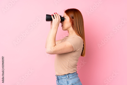 Young redhead woman over isolated pink background with black binoculars