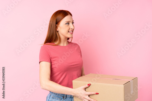 Young redhead woman over isolated pink background holding a box to move it to another site © luismolinero