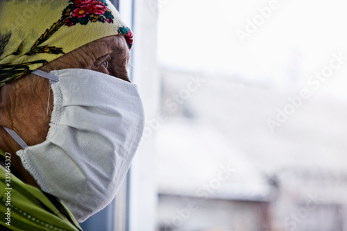 An old grandmother misses the street during quarantine due to an outbreak of coronary virus infection. A masked grandmother is looking through the glass at the street. Stay at home. Be safe on self-is