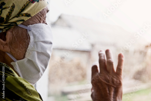 An old grandmother in a medical mask looks through the window into the street. An elderly woman in a mask on her face on self-isolation during quarantine due to a coronavirus pandemic