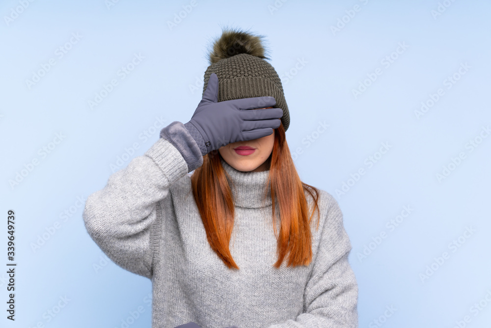Young redhead Russian woman with winter hat over isolated blue background covering eyes by hands