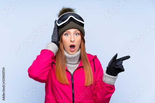 Skier redhead woman with snowboarding glasses over isolated blue wall surprised and pointing finger to the side