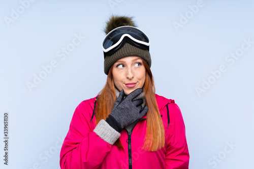 Skier redhead woman with snowboarding glasses over isolated blue wall thinking an idea © luismolinero