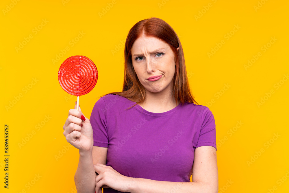 Young redhead woman holding a lollipop over yellow background with sad expression Young redhead woman holding a lollipop over yellow background with sad expression