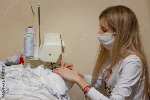 blonde girl with a white mask on her face sews gauze protective masks on an overlock sewing machine to protect against coronavirus during an epidemic photo