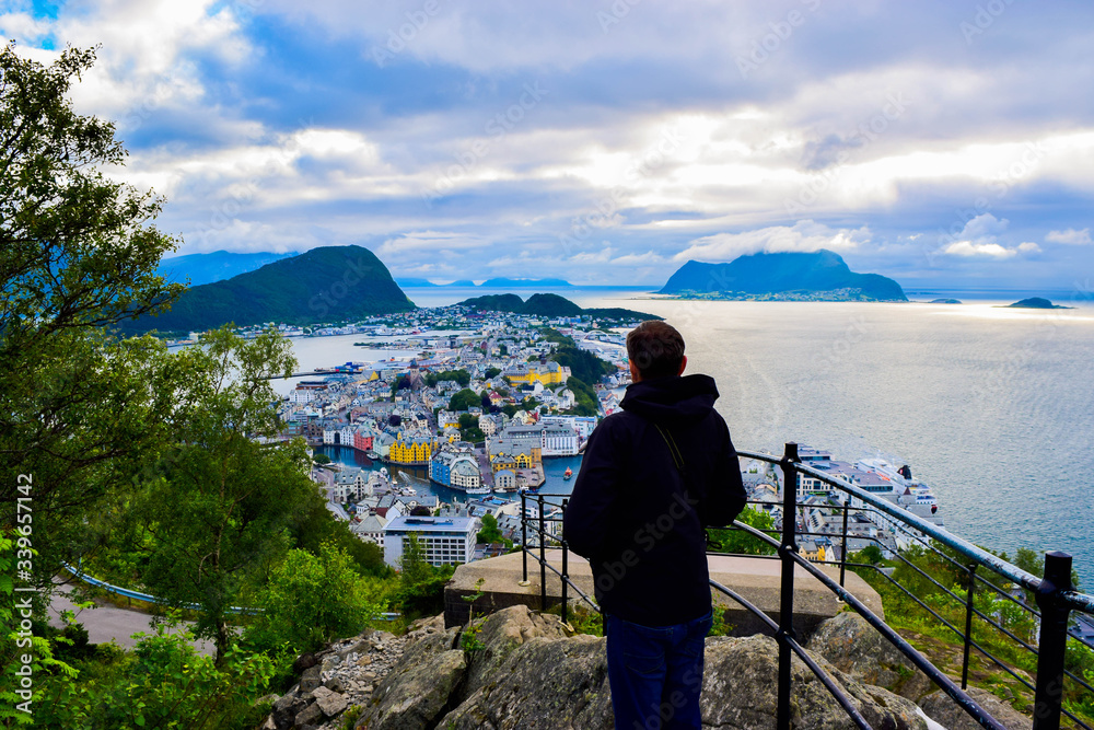 Tourist man looks on the Alesund cityscape from Aksla viewpoint. Neoclassical and neo-Gothic stone buildings. Art Nouveau architecture. Sun rays illuminate Atlantic Ocean and islands. Norway.