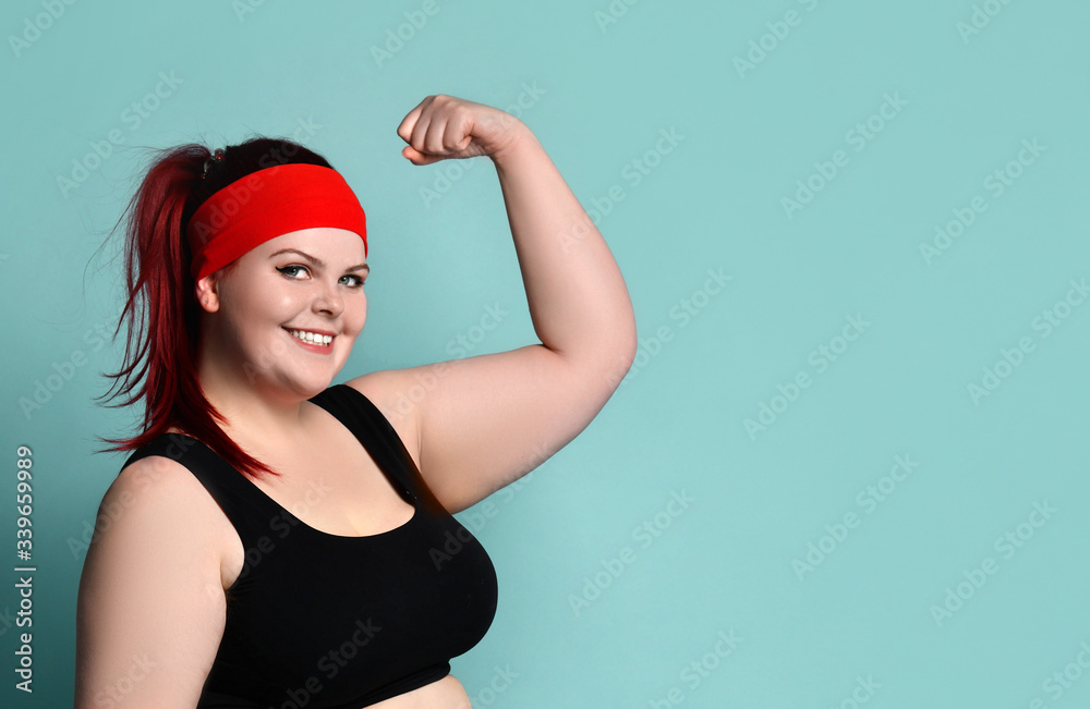 Portrait of a young fat ginger model in a red headband, black top. She  shows her strength in the arms of her biceps. Stock Photo | Adobe Stock