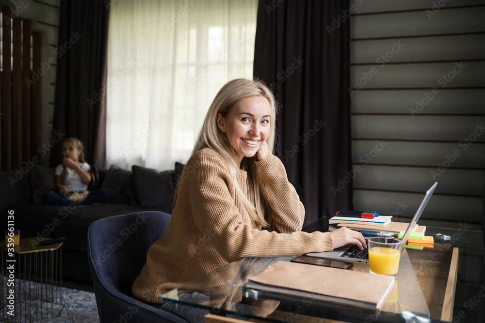 Beautiful blonde woman looking at camera and smiling while working on laptop computer sitting at desk at home, her little daughter eating popcorn and watching tv on sofa in background - obrazy, fototapety, plakaty 
