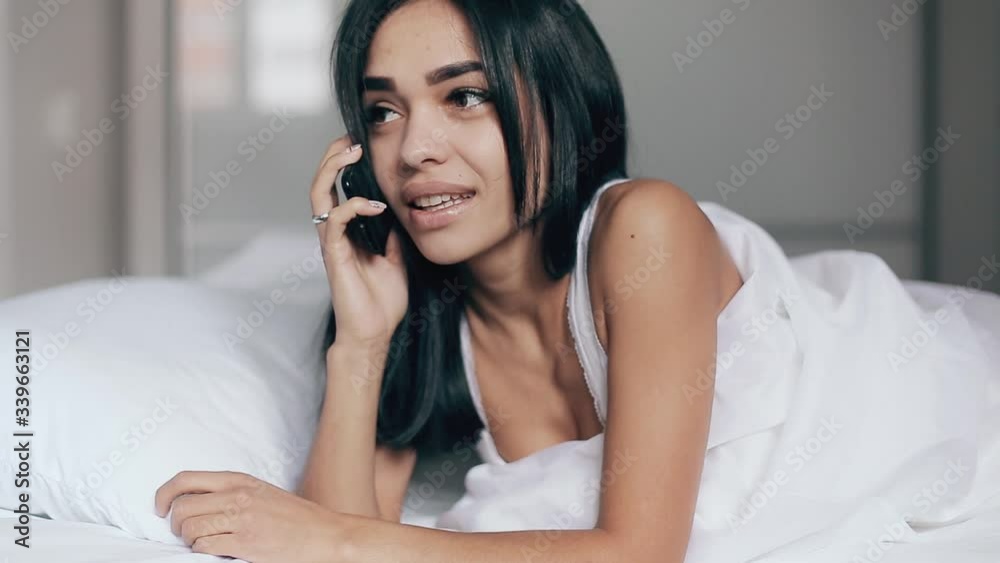Beautiful young brunette woman smiling lying talking smart mobile phone relax in the bedroom