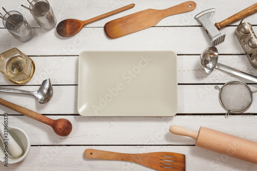 Prostokątna szklana biała miska leży na drewnianym stole. Na około porozkładane są stylowe naczynia kuchenne tworzące tło.