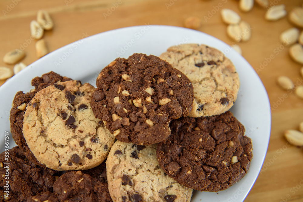 Close up photo with shallow depth of field of different cookies on a white oval shaped plate with peanuts around it on the table. Chocolate chip cookies and peanut chocolate cookies. 