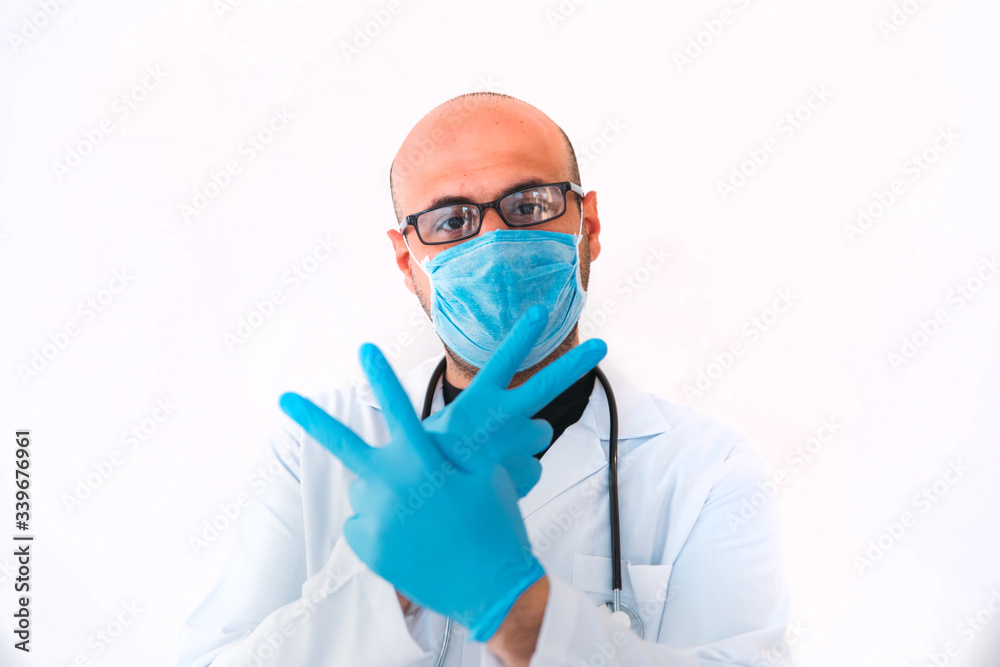 Portrait of a young male doctor making victory sign in medical mask, sterile gloves and stethoscope while looking at camera on isolated white.