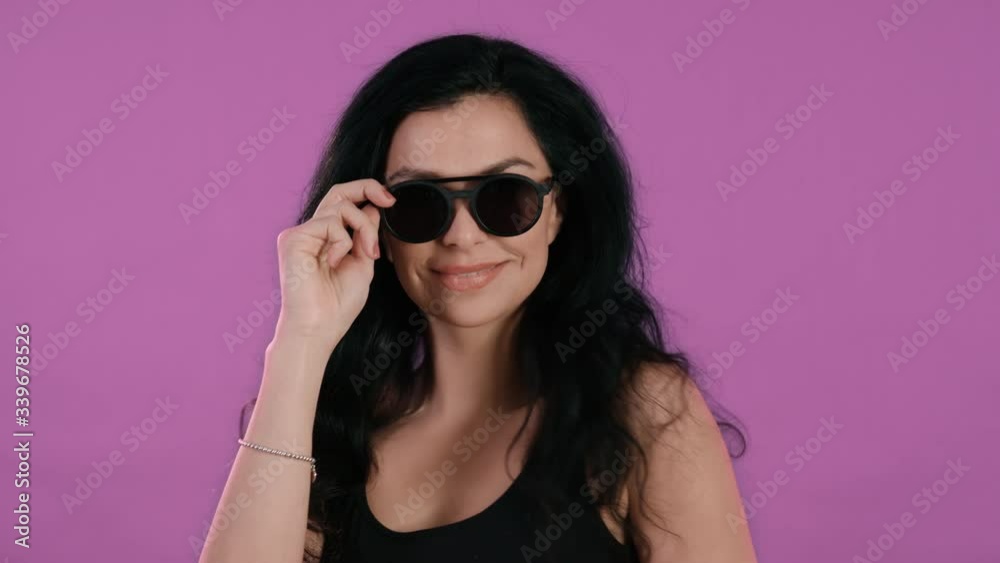 Portrait of beautiful young woman with black hair looking sexy over sun glasses in front of pink background shot in 4k super slow motion Portrait of beautiful young woman with black hair looking sexy over sun glasses in front of pink background shot in 4k super slow motion