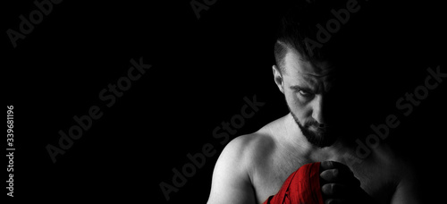 Banner image of a man’s fighter with a beard and red bandages on his hands. Head down, look from beneath the forehead. Black white image with red. Place for text. Black background.