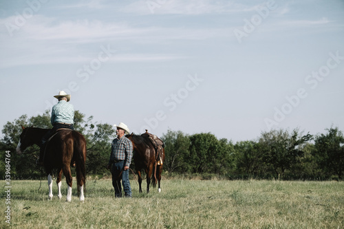 Western lifestyle shows cowgirl and cowboy in rural Texas field with horses. Copy space on sky background of farm.