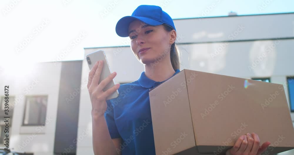 View from below on beautiful young Caucasian female postal worker in blue cap and with carton box tapping on smartphone outdoors. Pretty girl texting sms and scrolling on mobile phone at street.