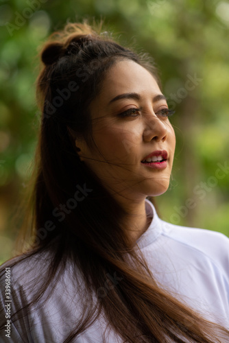 Smiling asian woman in park looking away from camera. Happy asia female black long hair in white shirt on green bokeh background.