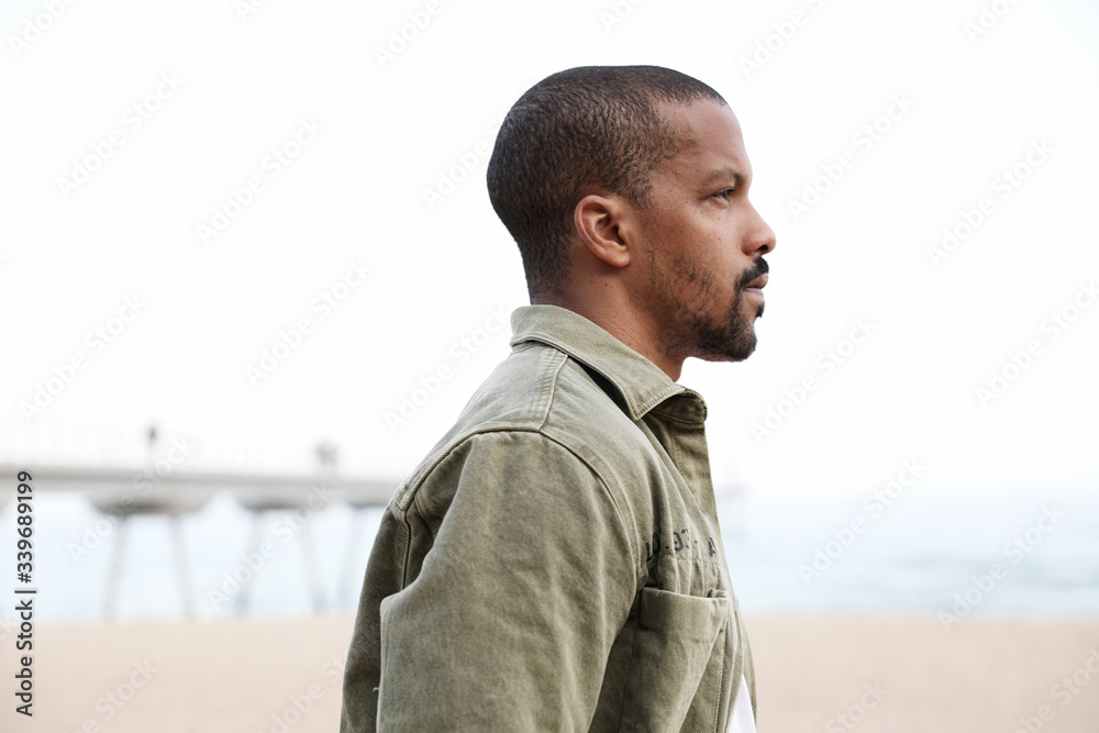 Fototapeta premium Side portrait of black young man walking on beach towards water. Outdoor.