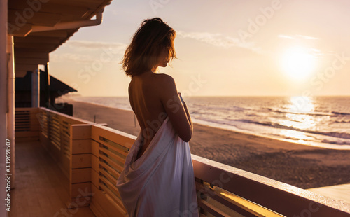 a half-naked girl stands wrapped in a sheet with her back to the frame on the terrace. dawn by the sea