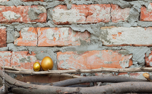 Two Golden Easter eggs stand on branches against a brick wall. Small and large painted eggs