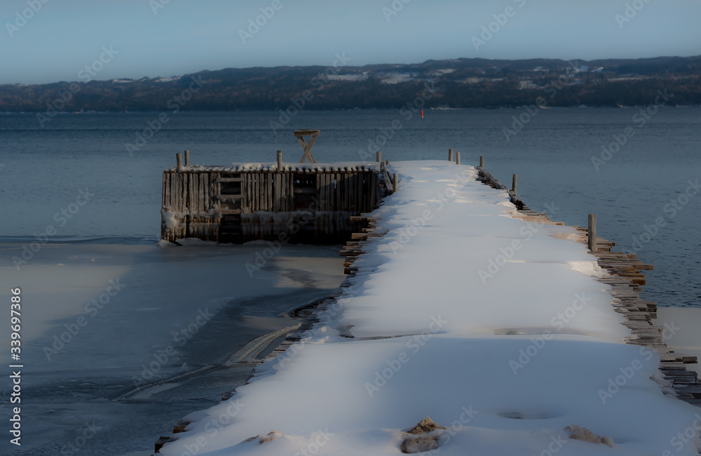An old worn and wooden wharf in an L-shape. The wharf extends into the cold Atlantic Ocean. There's snow on top of the wood pier. The background has mountains and smooth water.  