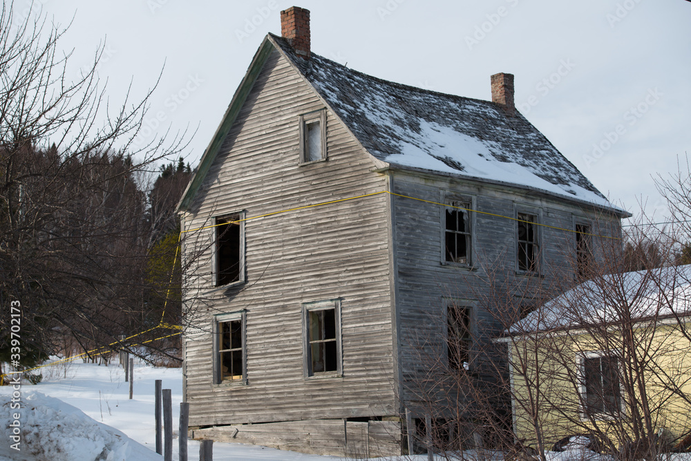 An abandoned two-storey scary building. The vintage weathered dilapidated residence has windows broken out, snow on the roof and grungy narrow grey clapboard on the exterior of the farmhouse.