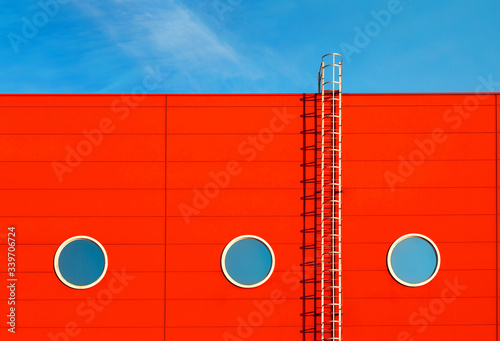 A fragment of a red modern facade of an industrial building, warehouse or shopping center with three round Windows and a metal staircase against the blue sky. Association with the ship's portholes