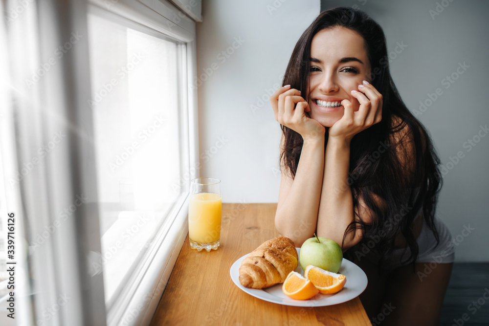 Good morning Young brunette woman in white underwear has breakfast on a wooden windowsill. A young female model eats. There is fresh orange or juice, croissant, fruit, apple on a white plate