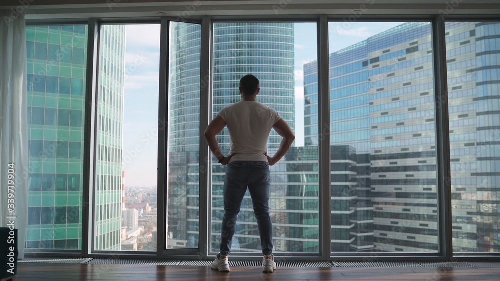 Fototapeta premium Athletic man looking out the window in the morning. Handsome young guy enjoying beautiful view on a sunny day. Rear back view Close up movement to the window