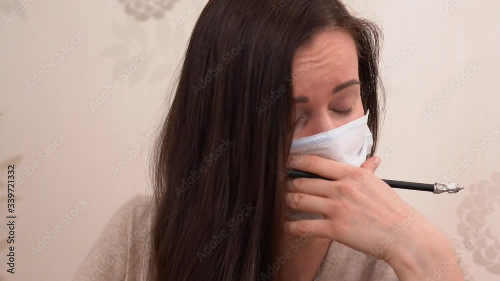 Portrait of sick Russian girl with long brown hair with black pencil in hand at home close up. Wonderful woman wear protective medical face mask, cough and feel pain in throat. Cover her mouth. Portrait of sick Russian girl with long brown hair with black pencil in hand at home close up. Wonderful woman wear protective medical face mask, cough and feel pain in throat. Cover her mouth.