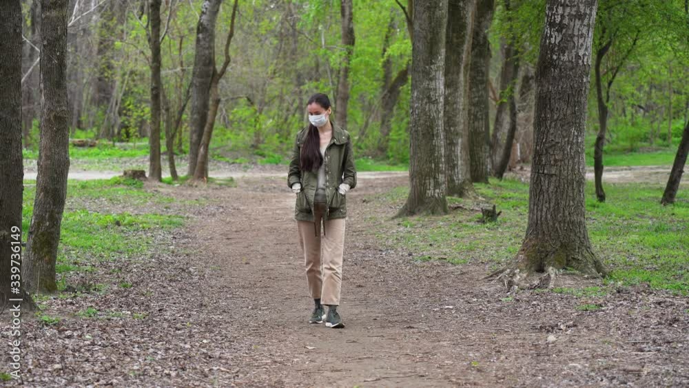 Young beautiful Caucasian woman with long dark brown hair in a blue protective medical mask walks alone in summer forest, among the trees, breathe fresh air in quarantine by pandemic of corona virus. Young beautiful Caucasian woman with long dark brown hair in a blue protective medical mask walks alone in summer forest, among the trees, breathe fresh air in quarantine by pandemic of corona virus.