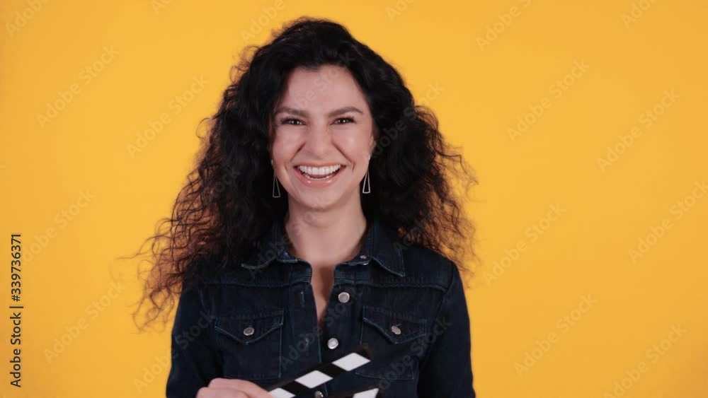 Portrait of beautiful young woman with black curly hair jumping twinking and clapping a clapperboard in front of yellow background shot in 4k Portrait of beautiful young woman with black curly hair jumping twinking and clapping a clapperboard in front of yellow background shot in 4k
