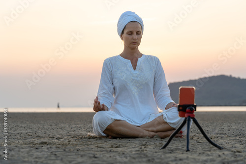 Young caucasian woman in white clothes and Indian turban meditating on a sandy tropical beach at sunrise and shoots himself on a smartphone