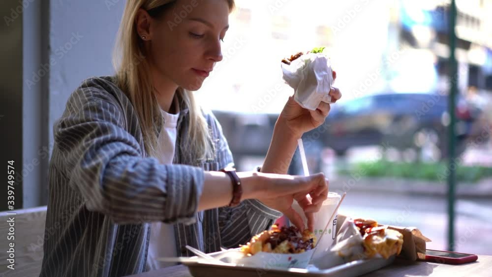 Happy hipster girl enjoying cheat meal day for eating unhealthy food after diet lifestyle tasting french fries and big hamburger, female gourmet drinking beverage during lunch
