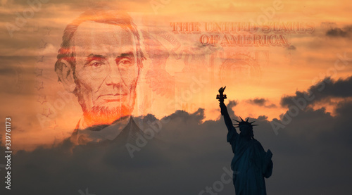 Statue of Liberty in silhouette with a portion of a warm-toned five-dollar bill in the horizon blending in with the sunrise
