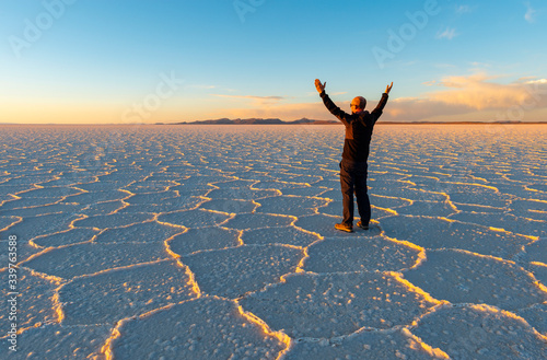 Sunset of the Uyuni Salt Flat Desert with Hexagon salt formations and a caucasian male tourist with stretched hands in the air, Bolivia.