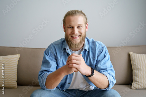Smiling millennial hipster guy blogger looking at camera video calling, recording vlog. Happy young man distance chatting at home office, streaming sitting on sofa. Headshot portrait. Webcam view