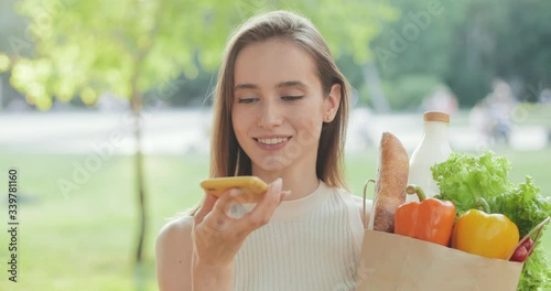 Young woman using voice recognition assistant to make request or record message on smartphone outdoors. Pretty girl talking on speakerphone while standing and holding shopping bag