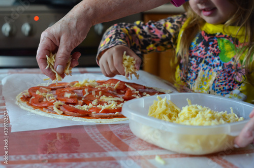 The hand of a woman and a child sprinkle grated cheese on an uncooked pizza. Joint cooking