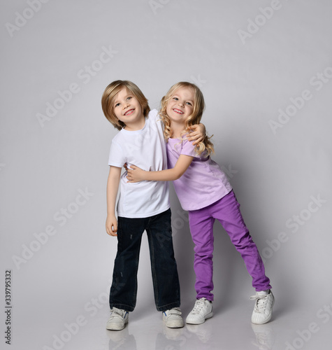 Two cheerful little blond kids girl and boy in stylish casual clothing have fun hugging smiling playing together