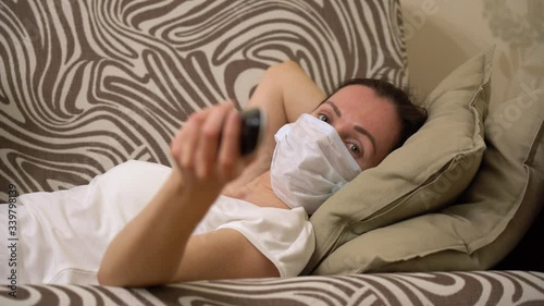 Alone beautiful lazy woman in white t-shirt, wear protective medical face mask, lying on sofa, holds a remote control, switches channels, watches TV. Stay isolation home self quarantine prevention