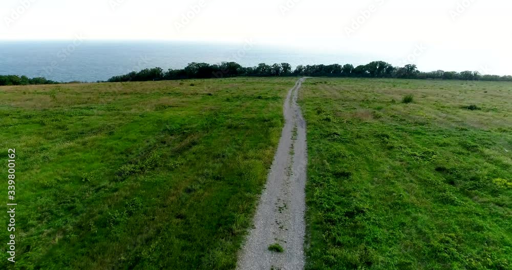 Flying over a dirt road that goes through a field with green grass towards the sea Flying over a dirt road that goes through a field with green grass towards the sea