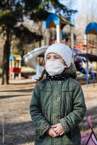 Sad little girl in a protective mask on the background of the closed playground outdoor. A child in a medical mask walks in the park. Quarantine measures against COVID-19. Coronavirus pandemic.