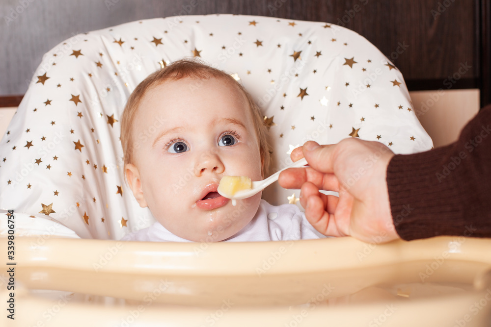 The child is funny, sitting on a highchair, scared. A one-year-old girl in a white sweater is sitting at a yellow table on a brown background, waiting for food with frightened big eyes. The child is funny, sitting on a highchair, scared. A one-year-old girl in a white sweater is sitting at a yellow table on a brown background, waiting for food with frightened big eyes.