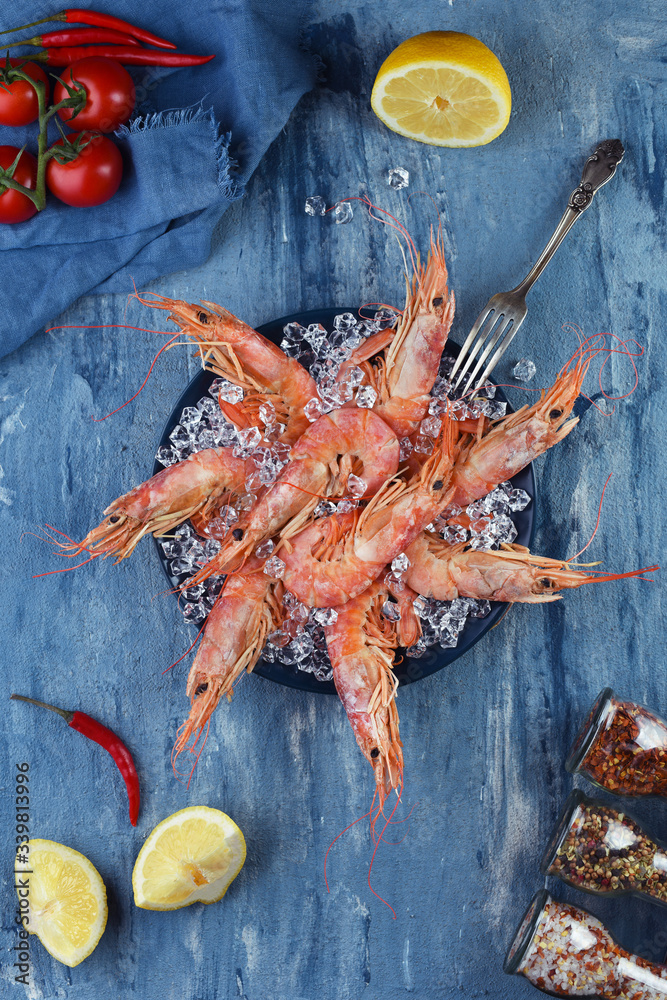 Set of king prawns in plate with fork. Ice in blue plate with shrimps and vegetables around. Sea food ready to eat. Table with seafood top view. Blue background. Set of king prawns in plate with fork. Ice in blue plate with shrimps and vegetables around. Sea food ready to eat. Table with seafood top view. Blue background.