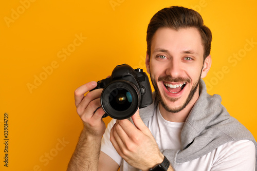 A happy photographer in white t-shirt with a beige sweater on his shoulders, has a black apple watch on, laughing, looks at the camera, holding the camera with both hands, has fun