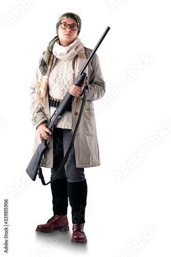 A full-length woman in a light coat, with a scarf on her head, with a gun in her hands and a bandoleer on her belt, poses on a white background. The woman is a hunter.