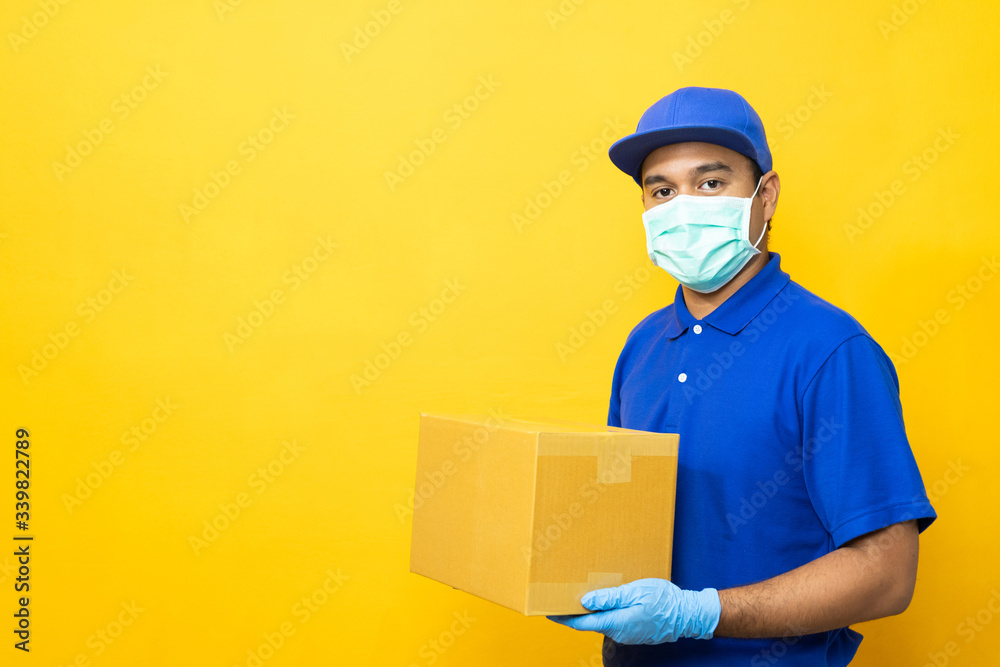 Delivery man blue uniform wearing rubber gloves and mask holding parcel cardboard box on yellow background. Delivery man blue uniform wearing rubber gloves and mask holding parcel cardboard box on yellow background.