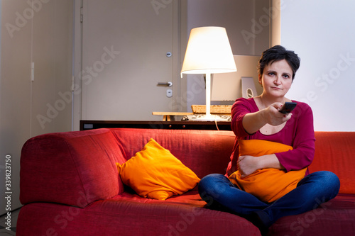 Donna Bianca con i capelli scuri, vestita casual, abbracci il cuscino seduta nel divano  mentre cambia canale alla tv con il telecomando photo